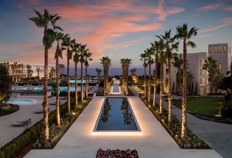 Palmengesäumter Pool des Ikos Resorts bei Sonnenuntergang mit beleuchtetem Gehweg