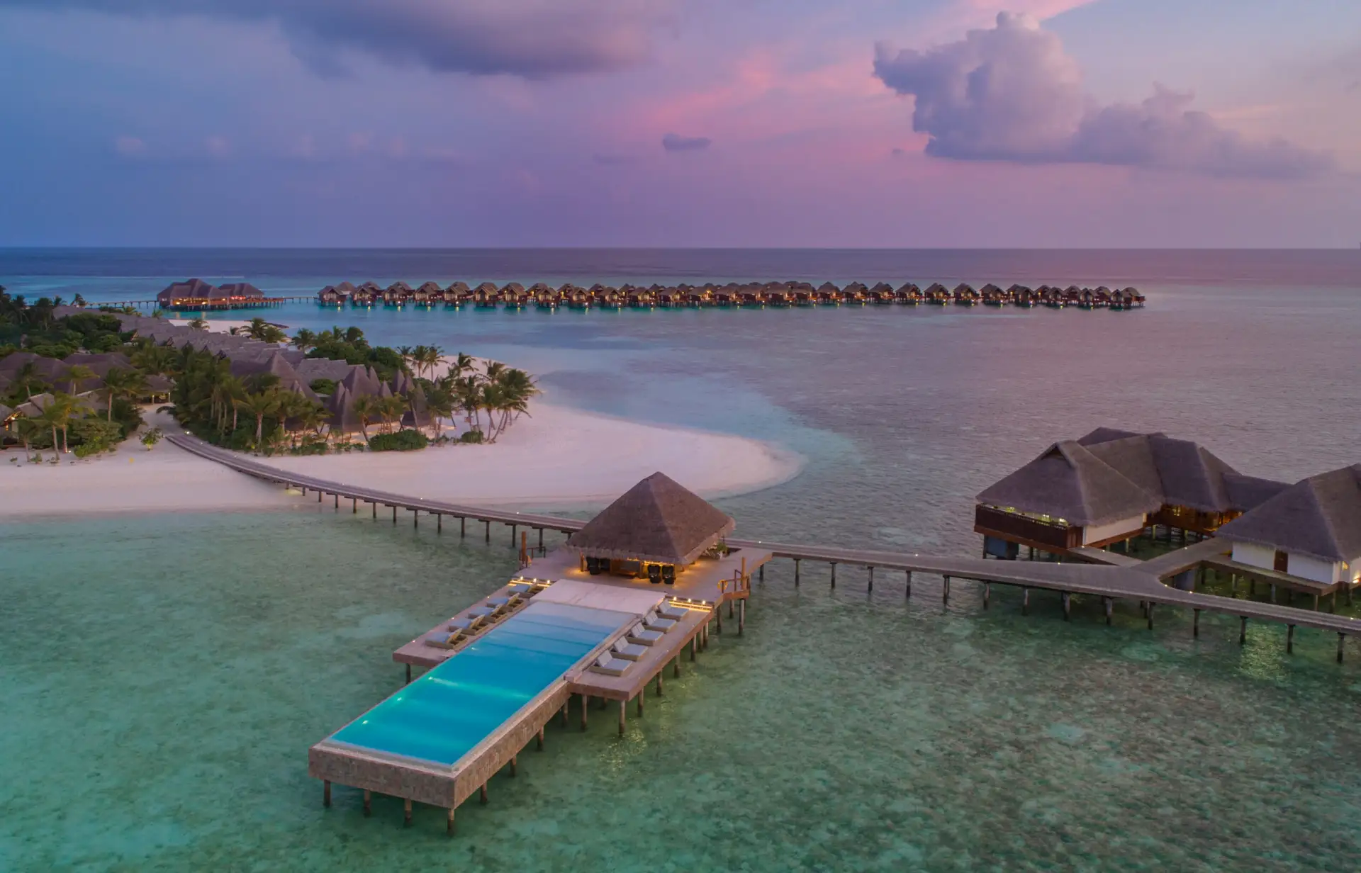 Infinity-Pool im Sonnenuntergang mit Sicht auf eine tropische Insel und Villen über dem Meer auf den Malediven